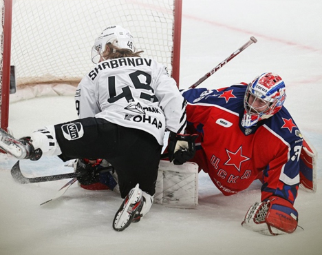 На изображении хоккейный вратарь в синей форме защищает ворота от игрока в бело-черной форме, который падает перед воротами. Ситуация напряженная, игроки сосредоточены на шайбе