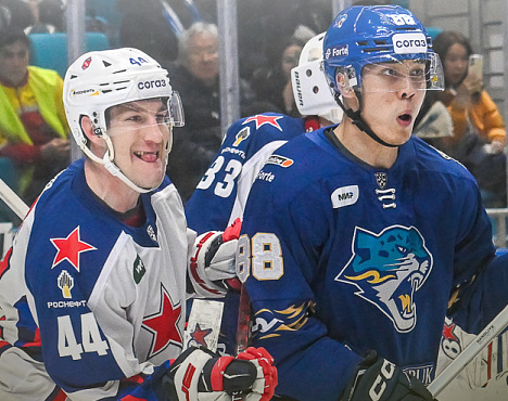 Хоккеисты в спортивной форме на льду во время игры. Один в синей форме, другой в белой с красными и синими элементами. Оба активно реагируют на происходящее. На заднем плане размытого общения