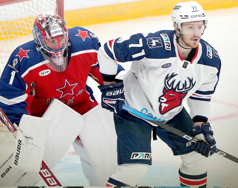 Хоккейный вратарь в красной форме защищает ворота. Рядом игрок в белой форме с логотипом оленя на груди. Оба активно участвуют в игре. На льду виден логотип Париматч