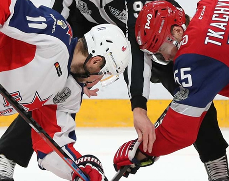 На изображении два хоккеиста в спортивной форме наклонены, готовясь к вбрасыванию шайбы. Они носят шлемы и защитное снаряжение. Рядом судья в черно-белом. Ледовый каток и желтая разметка видны на заднем плане