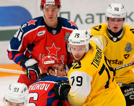 На изображении хоккеисты в форме красного и жёлтого цвета активно участвуют в игре на льду. Двое из них носят синие шлемы, остальные - белые. Видно напряженное противостояние между командами