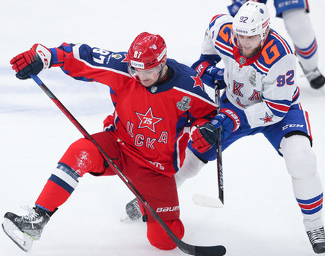 Хоккеисты в борьбе за шайбу на льду. Игрок в красной форме с номером 87 противостоит сопернику в бело-синей форме с номером 92. Обе команды активно соревнуются в динамичном моменте игры