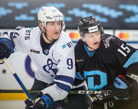 На изображении двое хоккеистов в игре. Один в белой форме с номером 94, другой в тёмной форме с номером 15. Они соревнуются за шайбу на льду. Видны логотипы на форме и шлемах игроков