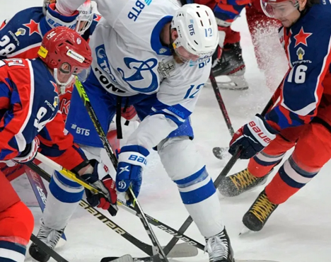 Хоккейный матч: игроки в форме сине-белой и красной расцветки соревнуются за шайбу на льду. Действие фокусируется на напряженной борьбе у коньков в центре кадра