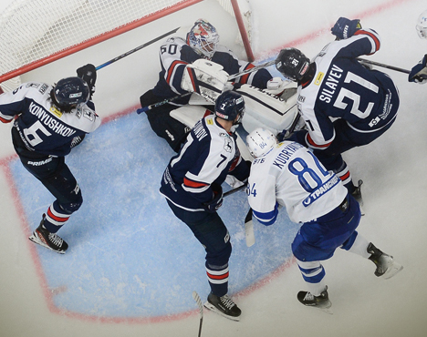 Хоккейный матч: группа игроков в тёмной и бело-голубой форме борется за шайбу возле ворот. Вратарь в белой форме готов отразить атаку. Игра проходит на ледовой площадке с видимой разметкой зоны ворот