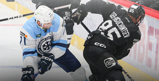 На изображении два хоккеиста в моменте игры. Хоккеист в форме светлых тонов пытается отобрать шайбу у игрока в черной форме. Они находятся на льду возле бортика, интенсивно сражаясь за владение шайбой