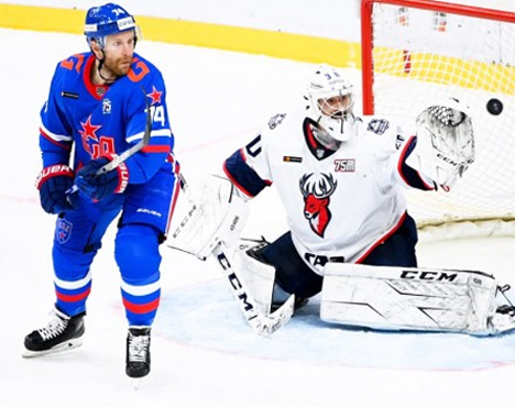 Хоккеист в синей форме стоит рядом с вратарём в белом, который ловит шайбу. На льду логотипы команд, на заднем плане видна хоккейная сетка