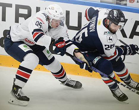 На изображении два хоккеиста в экипировке соперничают за шайбу на льду возле борта. Один из игроков в бело-синей форме, другой — в тёмно-синей с красными элементами. Игра проходит на арене