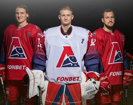 Три хоккеиста в форме клуба «Локомотив». В центре вратарь в белой форме, по бокам два игрока в красной. Все стоят на тёмном фоне, держа хоккейное снаряжение. Логотип команды на форме