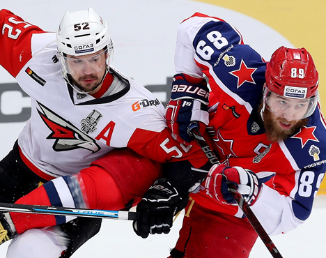 На изображении два хоккеиста в игровой борьбе. Спортсмен в красно-белой форме противостоит игроку в красно-синей форме. Оба держат клюшки, активно взаимодействуя на льду