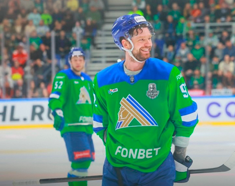 Хоккеисты на льду во время игры. Игрок в зеленой форме с номером 92 улыбается, держа клюшку. На заднем плане игрок с номером 52 и размытые зрители на трибунах